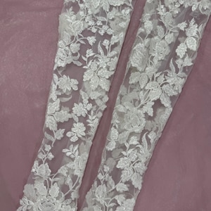 May include: Two white lace sleeves with floral embroidery. The delicate fabric features detailed patterns of flowers and leaves. The sleeves are displayed against a soft, mauve background, showcasing the intricate design and texture of the lace.