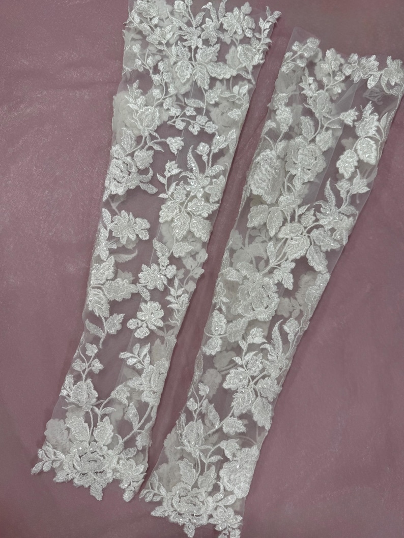 May include: Two white lace sleeves with floral embroidery. The delicate fabric features detailed patterns of flowers and leaves. The sleeves are displayed against a soft, mauve background, showcasing the intricate design and texture of the lace.