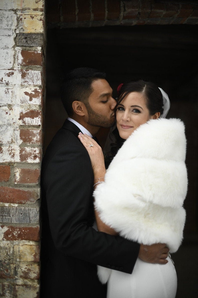 May include: A couple poses for a wedding photo. The groom is wearing a black suit and the bride is wearing a white dress with a white fur shawl.