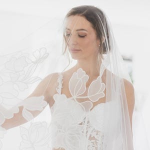 Op de afbeelding: Een witte bruids sluier met delicate bloemen borduurwerk. De sluier is doorschijnend en heeft een zachte, vloeiende textuur.