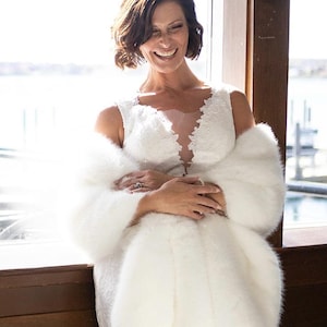 May include: A smiling person wearing a white wedding dress with a lace bodice and a faux fur wrap. The dress has a plunging neckline. The wrap is white and fluffy. The background is a window with a view of the water.