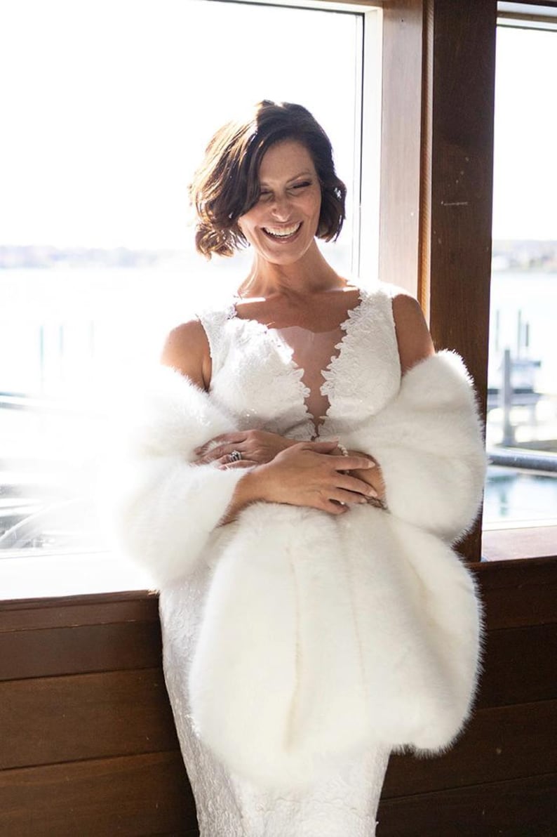 May include: A smiling person wearing a white wedding dress with a lace bodice and a faux fur wrap. The dress has a plunging neckline. The wrap is white and fluffy. The background is a window with a view of the water.