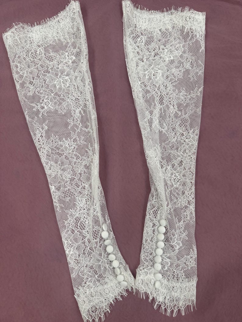 May include: Two white lace sleeves with floral patterns and scalloped edges. Each sleeve has a row of small, round buttons. The sleeves are made of delicate, sheer lace.