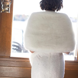 May include: A white faux fur shawl draped over a white lace dress. The shawl is fluffy and covers the shoulders. The dress is fitted and has a detailed lace pattern. The scene is set near a window with natural light.