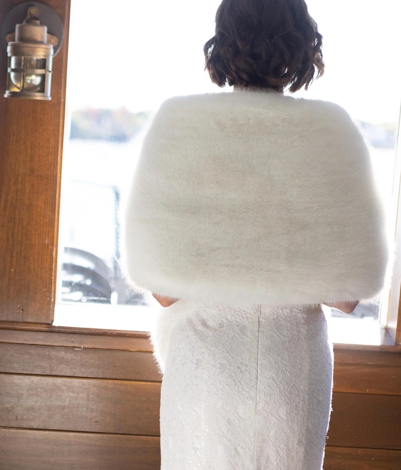 May include: A white faux fur shawl draped over a white lace dress. The shawl is fluffy and covers the shoulders. The dress is fitted and has a detailed lace pattern. The scene is set near a window with natural light.