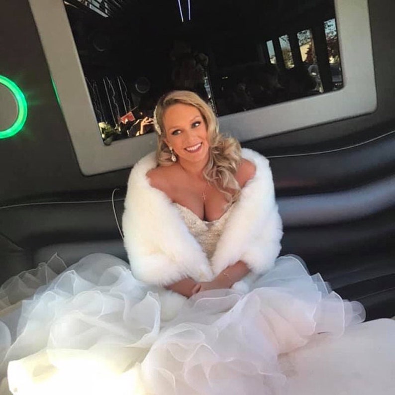 May include: A bride in a white wedding dress with a white fur shawl, sitting in a black limousine.