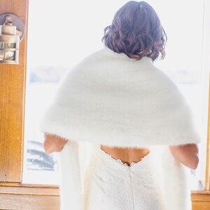 May include: A white faux fur shawl draped over a white lace dress. The shawl is fluffy and covers the shoulders. The dress has a low back and a delicate lace pattern. The scene is illuminated by natural light from a window.