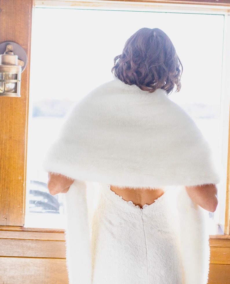 May include: A white faux fur shawl draped over a white lace dress. The shawl is fluffy and covers the shoulders. The dress has a low back and a delicate lace pattern. The scene is illuminated by natural light from a window.
