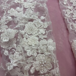 May include: Close-up of white floral lace fabric with intricate detailing. The lace features delicate flower patterns with beaded accents, set against a soft pink background. The design is elegant and detailed, suitable for bridal or formal wear.