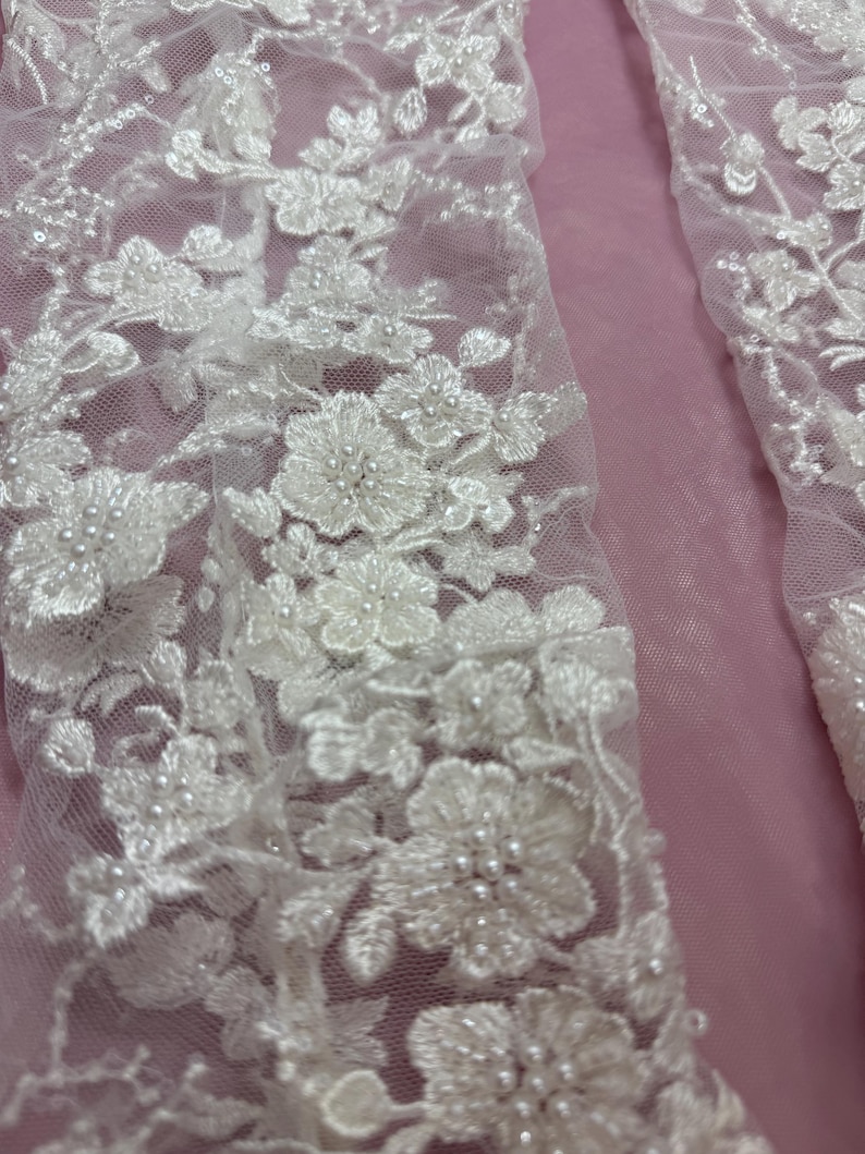 May include: Close-up of white floral lace fabric with intricate detailing. The lace features delicate flower patterns with beaded accents, set against a soft pink background. The design is elegant and detailed, suitable for bridal or formal wear.