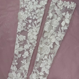 May include: Two white lace sleeves with floral embroidery. The sleeves are made of a sheer fabric and are decorated with detailed, raised floral patterns. The design includes various flower and leaf shapes, creating a delicate and elegant look.