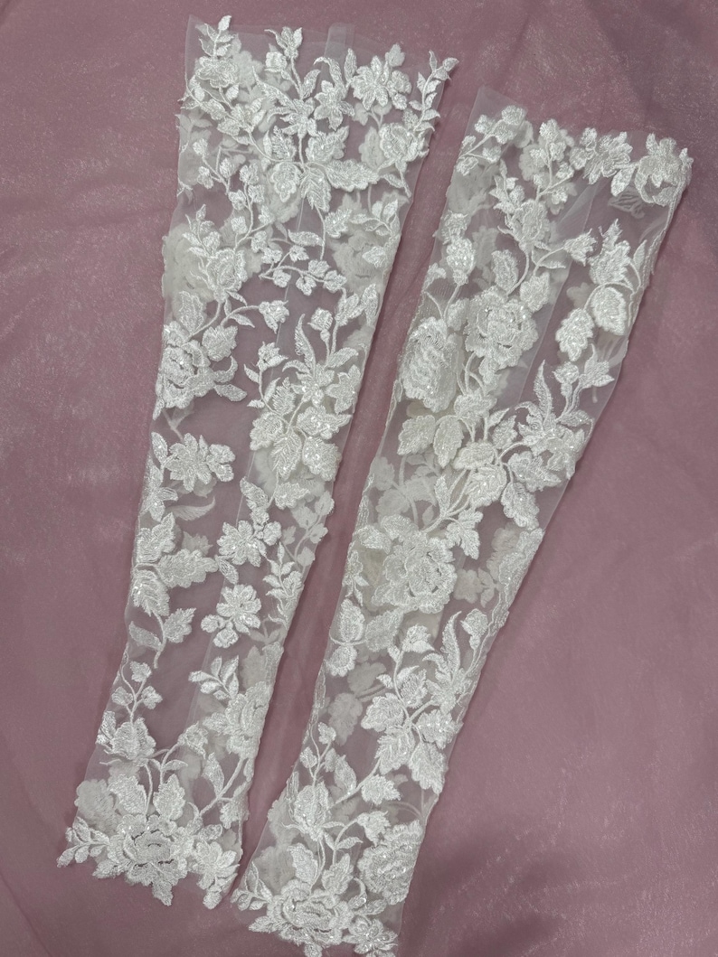 May include: Two white lace sleeves with floral embroidery. The sleeves are made of a sheer fabric and are decorated with detailed, raised floral patterns. The design includes various flower and leaf shapes, creating a delicate and elegant look.