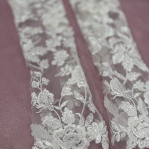 May include: Close-up of two white lace sleeves with floral embroidery. The delicate lace features detailed flower and leaf patterns, creating a romantic and elegant look. The sleeves are set against a soft, muted pink background, highlighting the intricate design.