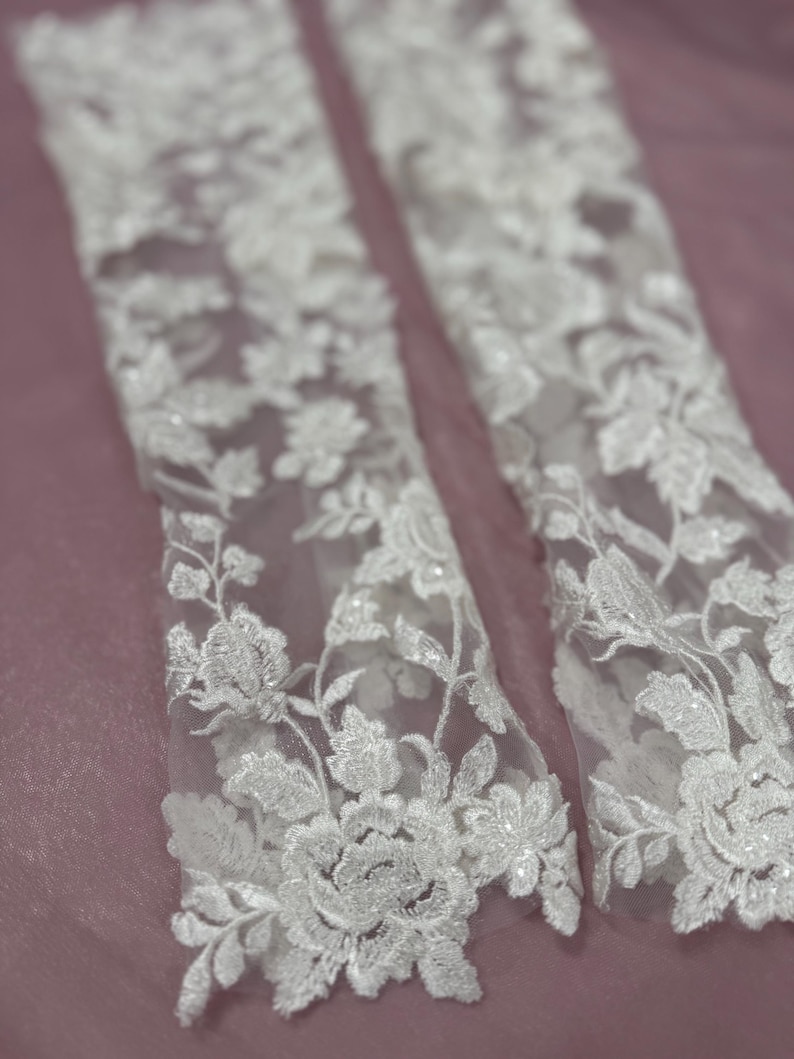 May include: Close-up of two white lace sleeves with floral embroidery. The delicate lace features detailed flower and leaf patterns, creating a romantic and elegant look. The sleeves are set against a soft, muted pink background, highlighting the intricate design.