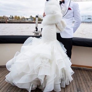 May include: A bride wearing a white wedding dress with a large, ruffled skirt and a white fur shawl. She is kissing the groom, who is wearing a white tuxedo with a black bow tie and a red pocket square.