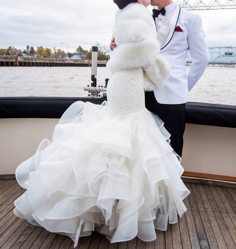 May include: A bride wearing a white wedding dress with a large, ruffled skirt and a white fur shawl. She is kissing the groom, who is wearing a white tuxedo with a black bow tie and a red pocket square.