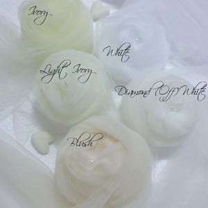 May include: Four fabric swatches in different shades of white: ivory, light ivory, diamond off white, and blush. Each swatch is gathered into a loose ball shape and has a small pearl bead in the center.