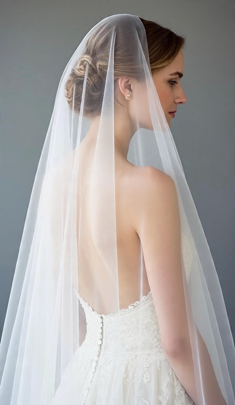 May include: A long, sheer white bridal veil made of fine tulle fabric. The veil is draped over the head and flows down the back, featuring a smooth, unadorned edge. It is worn over a white lace dress with a low back and a row of small fabric buttons.
