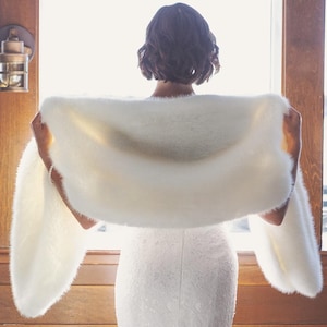 May include: A fluffy, off-white shawl is held up by a person, showcasing its soft texture. The shawl is draped over the shoulders, with the ends extending outwards. The background features a window and wooden accents, creating a warm, inviting atmosphere.