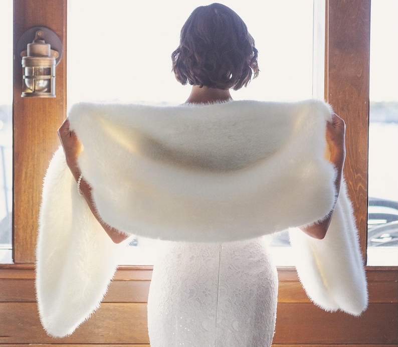 May include: A fluffy, off-white shawl is held up by a person, showcasing its soft texture. The shawl is draped over the shoulders, with the ends extending outwards. The background features a window and wooden accents, creating a warm, inviting atmosphere.
