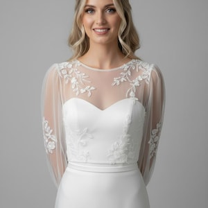 May include: A white bridal gown with a sweetheart neckline and a sheer, floral-embroidered overlay. The dress has long, sheer sleeves with matching floral details and a fitted bodice. The skirt is a simple, elegant design.