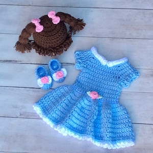 May include: A handmade crochet baby doll set in light blue, including a dress, shoes, and a doll head. The dress has white trim and a pink flower accent. The shoes are blue with white and pink flower details. The doll head is brown with pink bows.