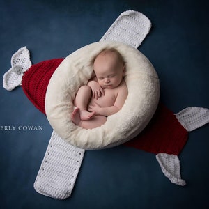 May include: A newborn baby sleeps peacefully inside a soft, white, circular nest, placed in the center of a red and white crocheted airplane. The airplane is set against a dark blue background. The image is a newborn photography concept.