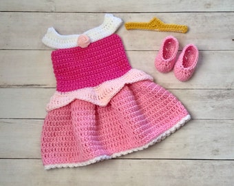 May include: A pink and white crocheted baby dress with a white crocheted flower on the bodice, a gold crown headband, and a pair of pink crocheted baby shoes.
