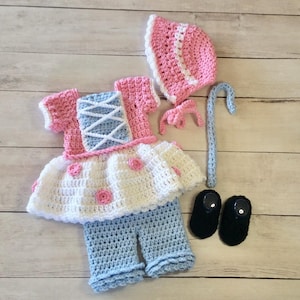 May include: A crochet baby outfit in a Bo Peep theme. The set includes a pink and white bonnet, a dress with pink accents and blue lacing, blue pants, a crochet shepherd's crook, and black booties. The outfit is handmade.