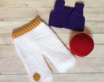 May include: A crocheted baby outfit in white, purple, and red. The outfit includes pants with a yellow trim, a vest, and a hat.