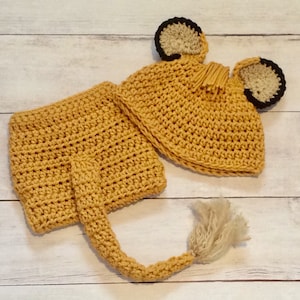 May include: A crocheted lion costume for a baby, featuring a hat with ears and a tail. The costume is made in a light brown color.