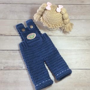 May include: A handmade crochet outfit, including blue overalls with a small patch and a blonde wig with pink bows. The overalls have white stitching on the cuffs and are secured with silver buttons.