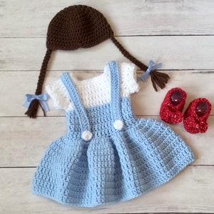 May include: A crocheted baby outfit in blue, white, and brown. The outfit includes a dress with a white bodice and blue skirt, a brown hat with pigtails, and red shoes.