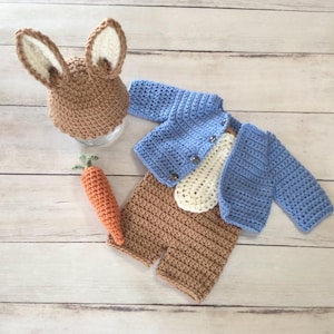 May include: A crochet baby outfit featuring a brown bunny hat with white inner ears, a light blue jacket, a cream-colored undershirt, and brown shorts. An orange carrot with green leaves completes the set. The items are arranged on a light wood surface.