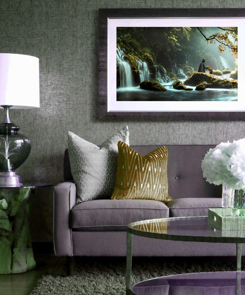 May include: A gray sofa with two throw pillows, one with a geometric pattern and the other with a gold chevron pattern. A framed picture of a waterfall in a forest hangs on the wall above the sofa. A glass coffee table sits in front of the sofa.