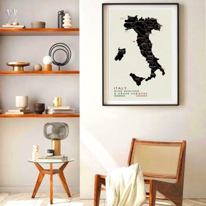 May include: A black and white map of Italy showing the wine regions and grape varieties. The map is framed and hanging on a white wall. A wooden chair with a white throw blanket is in the foreground.