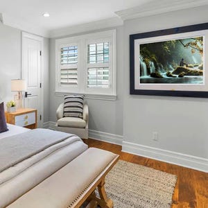 May include: A white bed with a gray blanket and a wooden bench at the foot of the bed. There is a white nightstand with a lamp and a small plant on it. A white chair with a gray and white striped cushion is in the corner of the room. A framed picture of a waterfall in a forest is hanging on the wall.