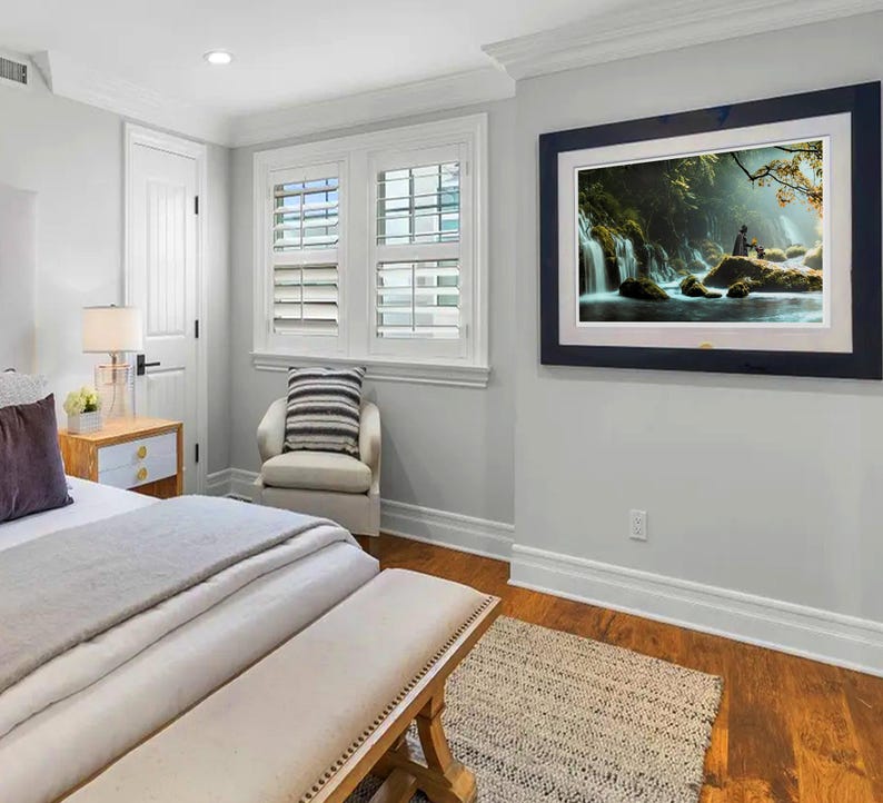 May include: A white bed with a gray blanket and a wooden bench at the foot of the bed. There is a white nightstand with a lamp and a small plant on it. A white chair with a gray and white striped cushion is in the corner of the room. A framed picture of a waterfall in a forest is hanging on the wall.