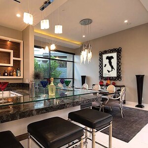 May include: A modern kitchen with a bar area featuring a granite countertop and black leather bar stools. The bar area has a glass top and a built-in liquor cabinet. The kitchen also has a dining area with a glass table and chairs. The dining area has a large black and white framed print of a map of Italy on the wall.