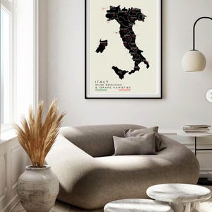 May include: A black and white map of Italy showing the wine regions and grape varieties. The map is framed and hanging on a white wall above a beige couch with a brown pillow. There is a vase with dried pampas grass in front of the couch.