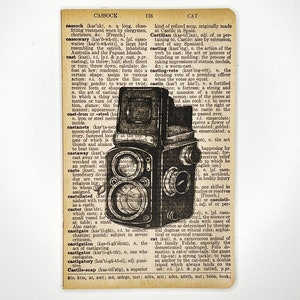 May include: A vintage camera with a black and silver body and a lens with a black leather covering. The camera is sitting on a page from a dictionary with the word "cast" highlighted.
