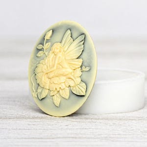 May include: Oval-shaped cameo with a fairy design in cream and pale green. The fairy is seated on a flower, with detailed wings and floral accents. The cameo is set against a white background.