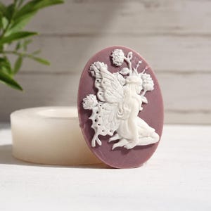 May include: Oval-shaped, mauve-colored soap mold featuring a white relief of a fairy with butterfly wings, flowers, and a flowing gown. A cylindrical, off-white mold is in the foreground. The fairy design is detailed and delicate.