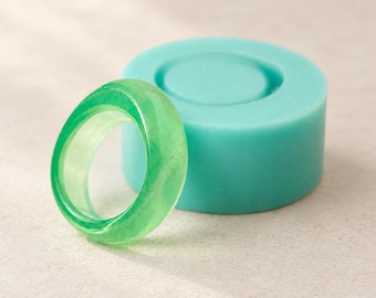 May include: A translucent, bright green resin ring with a smooth, polished finish positioned in front of a matching cylindrical silicone mold. The ring features a rounded band design, while the teal-colored mold has a circular indentation.