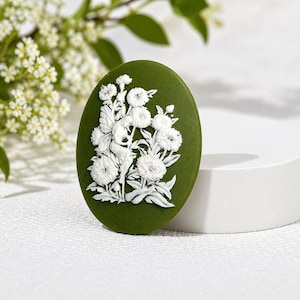 May include: An oval-shaped brooch featuring a matte olive green background with a raised white relief design of a figure standing among detailed floral blossoms and foliage. The piece has a smooth, flat surface and a distinct, high-contrast appearance.