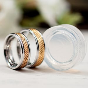 May include: Two wedding rings with a silver band and a gold textured centre band. A clear, round, glass object is next to the rings. The background is blurred with white flowers.