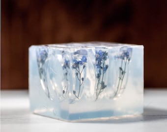 May include: Clear resin rings with embedded dried flowers. One ring features a large yellow flower, the other has a blue flower. A clear resin block with blue flowers is also shown. The rings are on a white surface.