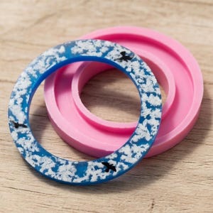 May include: A blue and white resin bangle bracelet with black bird accents, next to a pink silicone mould. The bracelet has a cloudy design. The background is a light brown wooden surface.