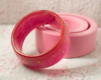May include: A vibrant red and pink bangle bracelet with gold flecks. The resin bracelet is set against a soft pink background. This bold, eye-catching accessory is perfect for adding a pop of color to any outfit.