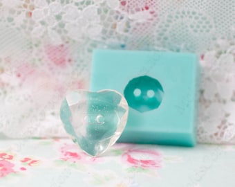 Pode incluir: Um botão transparente em forma de coração ao lado de um molde de silicone azul claro. O botão tem um design facetado e um pequeno orifício. O molde é quadrado com uma reentrância em forma de botão.